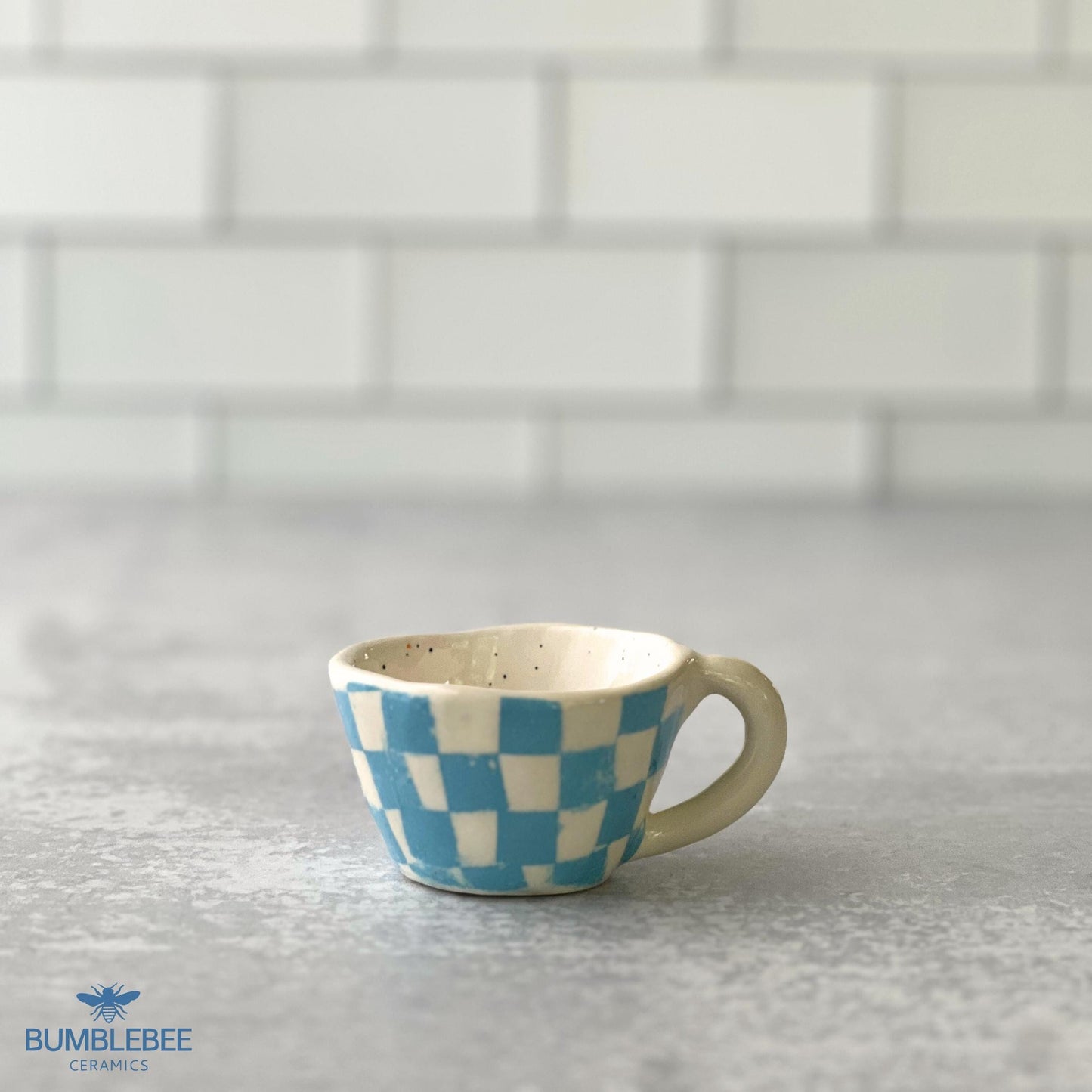 A small handmade espresso cup with a blue and white checkerboard pattern is placed on a gray countertop. The backdrop features clean white subway tiles, and the Bumblebee Ceramics logo is displayed in the corner.
