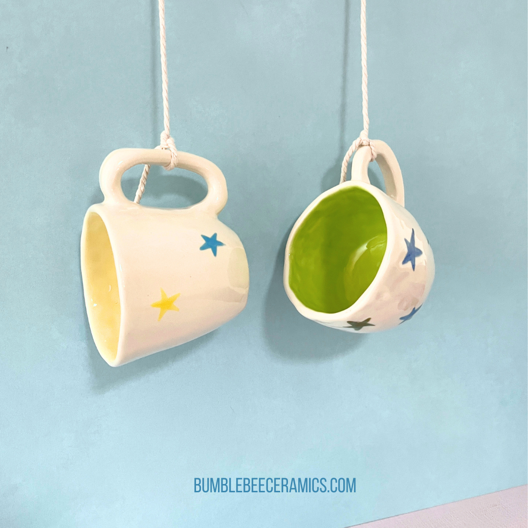 Two handmade ceramic espresso cups with colorful star designs hang by their handles from white cords against a soft blue background. One cup has a yellow interior and the other features a bright green interior, showcasing their whimsical and playful design.