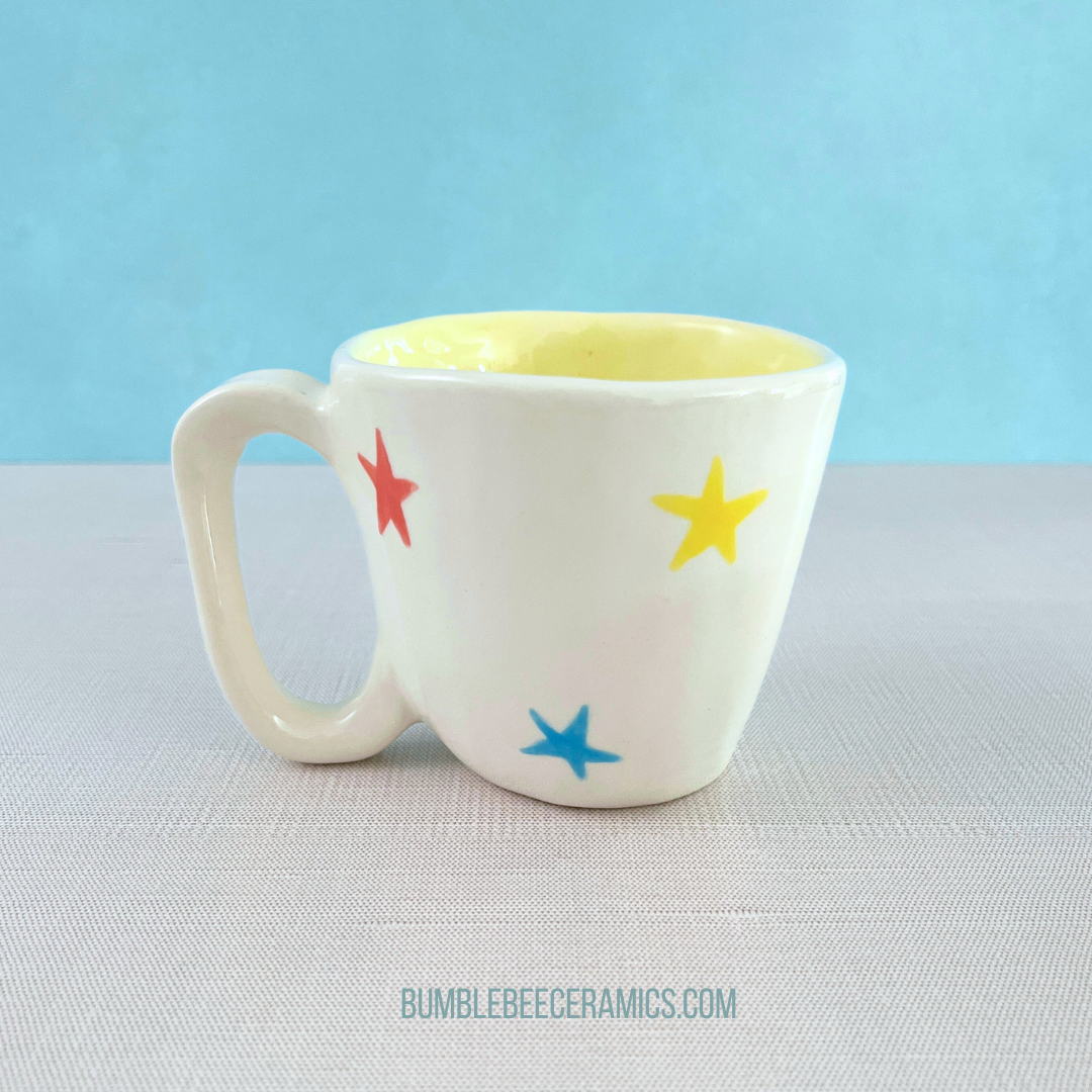 A white handmade ceramic mug with a yellow interior and colorful star designs in red, yellow, and blue on the outside. The mug is placed on a light surface with a soft blue background, and the text "BUMBLEBEECERAMICS.COM" is visible below.