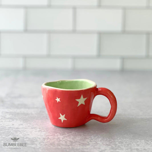 A bright coral handmade ceramic cappuccino cup with white stars and a soft green interior rests on a gray surface in front of a white tile background. It has a rounded, organic shape with a playful finish.