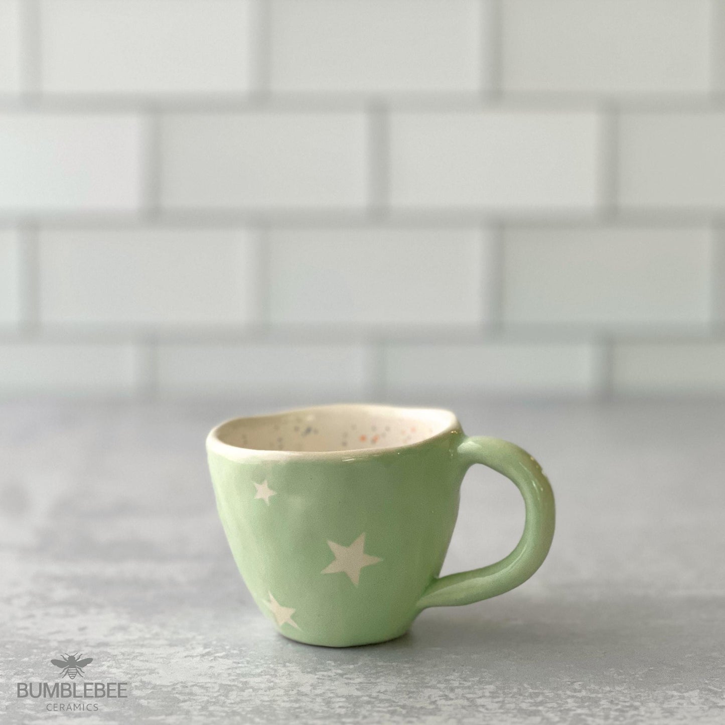 A handmade ceramic cappuccino cup with a misty green exterior and white star pattern sits on a gray countertop in front of a white tile backsplash. The interior features delicate colored speckled details.