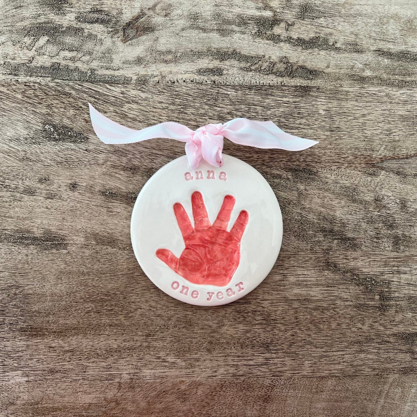 Round ceramic ornament with a red baby handprint, personalized with the name Anna and the words 'one year,' tied with a light pink ribbon.