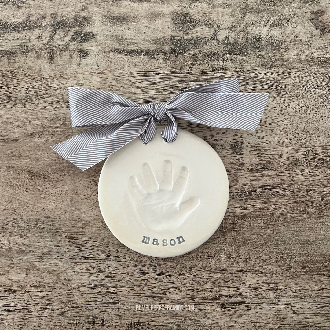 Round ceramic ornament featuring an impression of a baby handprint with the name Mason stamped below, finished with a gray herringbone ribbon.