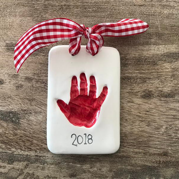 Decorative plaque with red baby handprint and '2018' on a wooden surface, tied with a red and white checkered ribbon.