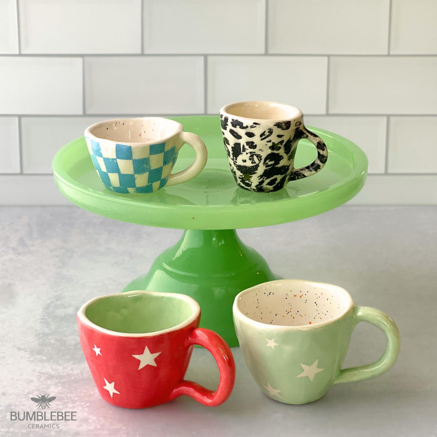 A vibrant display of four handmade ceramic espresso and cappuccino mugs arranged on a green cake stand. Designs include blue checkerboard, black-and-white leopard print, coral with stars, and misty green with white stars, all set against a white tiled background.