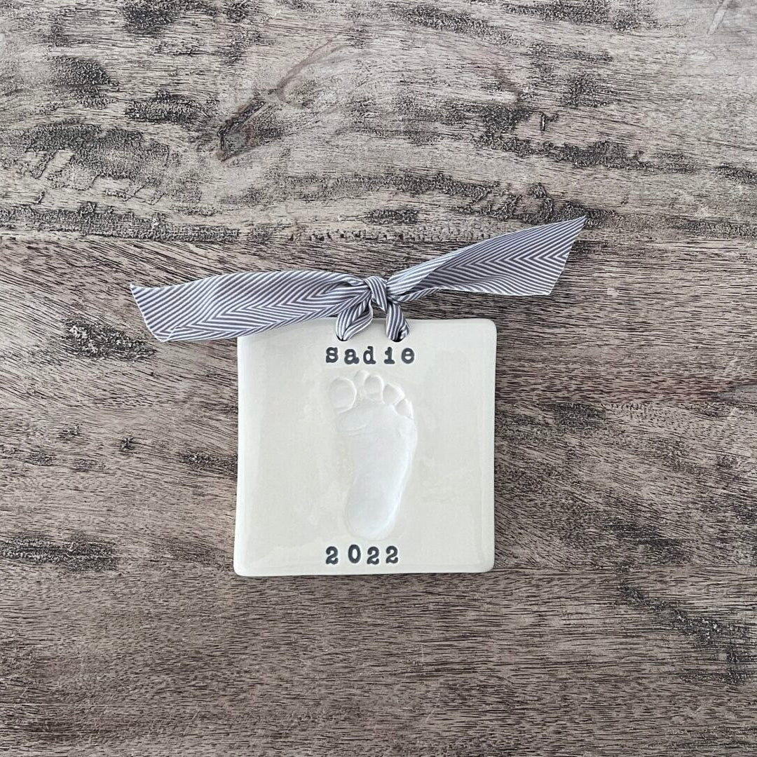 Rectangular ceramic keepsake with a white baby footprint, personalized with the name Sadie and year 2022, finished with a gray herringbone ribbon.
