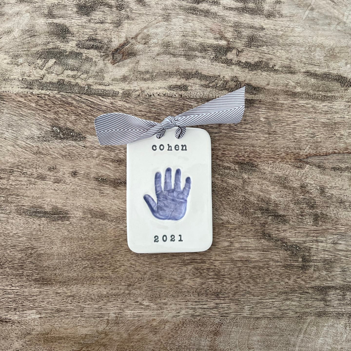 Rectangular ceramic keepsake with a purple baby handprint, personalized with the name Cohen and year 2021, finished with a gray herringbone ribbon.
