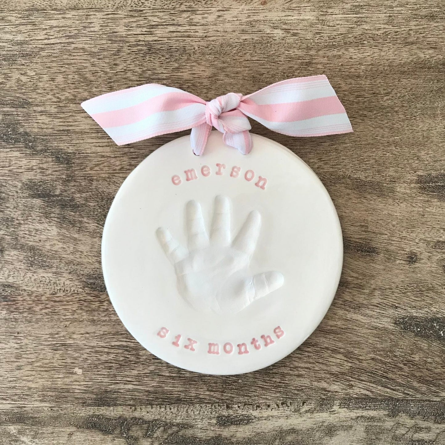 Round ceramic ornament featuring a white baby handprint with the name Emerson and the words 'six months,' tied with a pink and white striped ribbon.