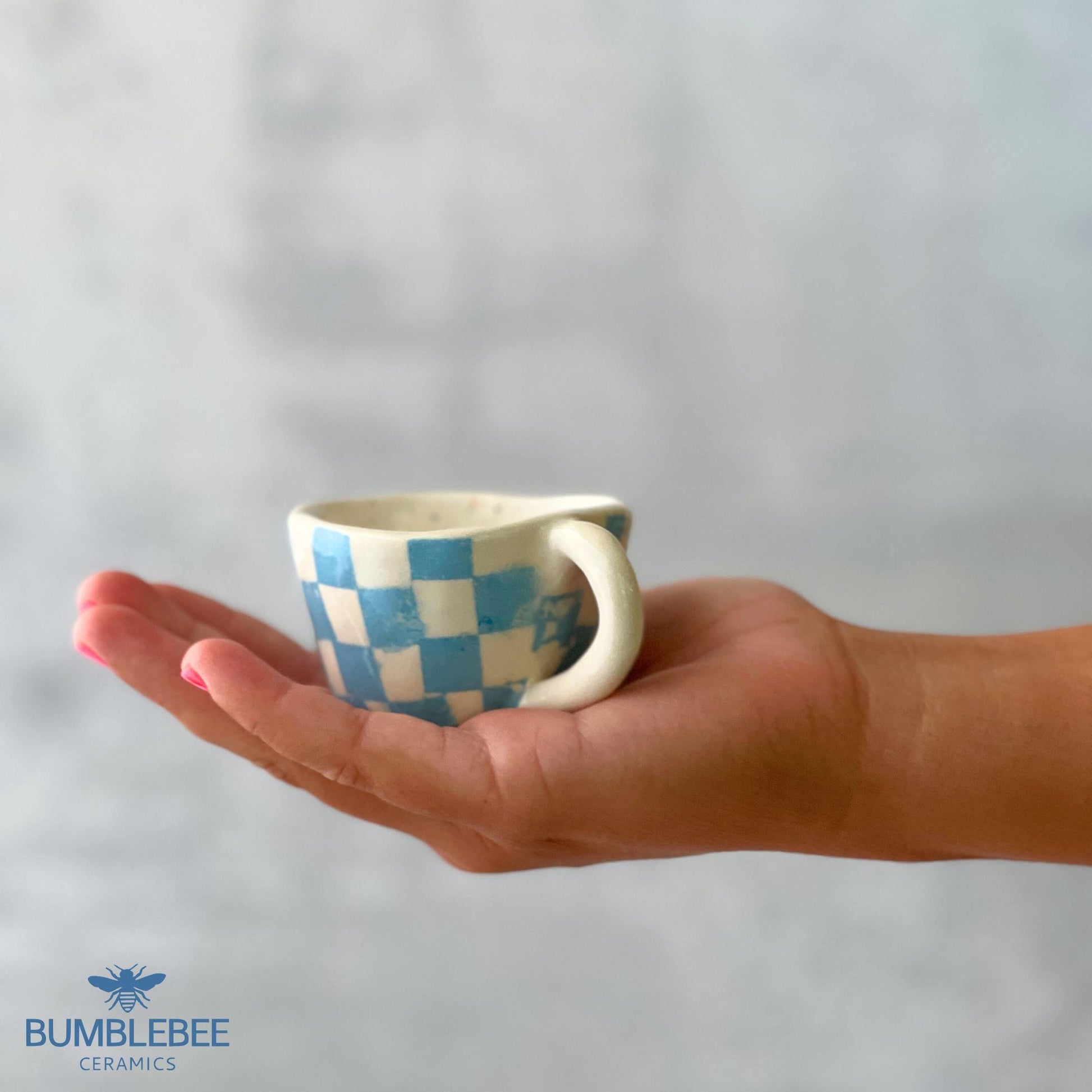 A tiny handmade ceramic mug with a blue and white checkerboard design rests gently in the open palm of a hand with pink painted nails. The image has a soft, neutral background and the Bumblebee Ceramics logo at the bottom.