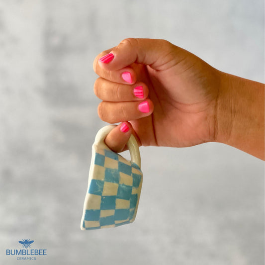 A small handmade ceramic espresso mug with a blue and white checkerboard pattern is held upright by a hand with bright pink painted nails. The background is softly blurred, keeping focus on the cup and hand.