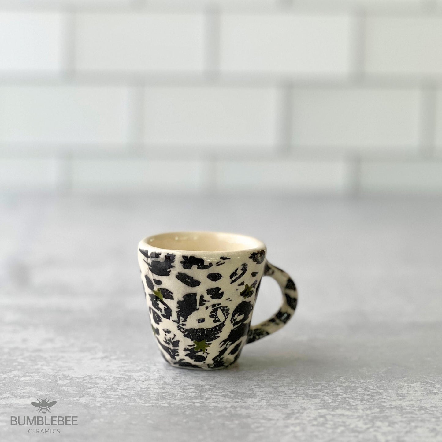 A handmade ceramic espresso cup featuring a bold black and white leopard print pattern sits on a gray countertop. The background is a clean white tile wall, and the Bumblebee Ceramics logo appears in the corner.