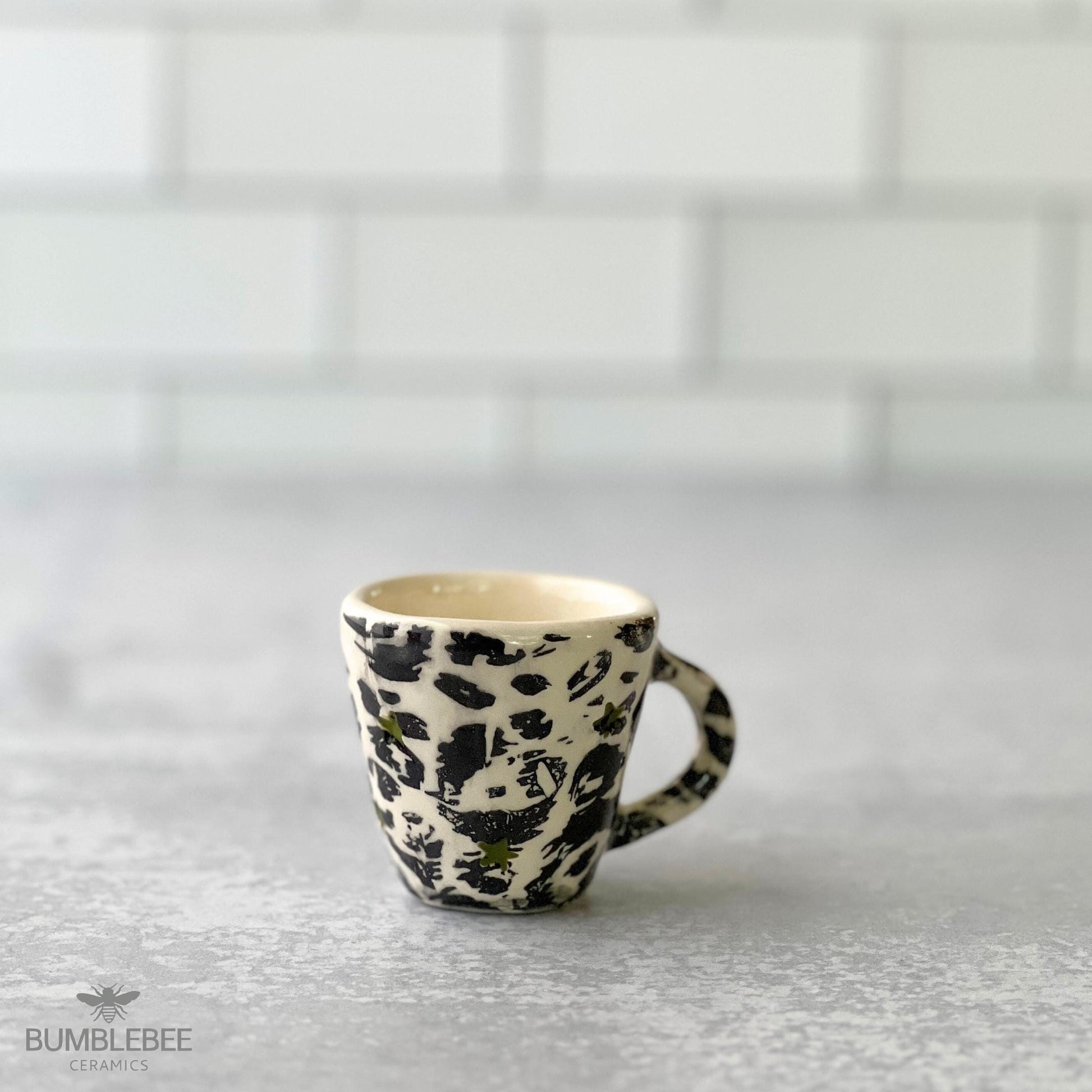 A handmade ceramic espresso cup featuring a bold black and white leopard print pattern sits on a gray countertop. The background is a clean white tile wall, and the Bumblebee Ceramics logo appears in the corner.