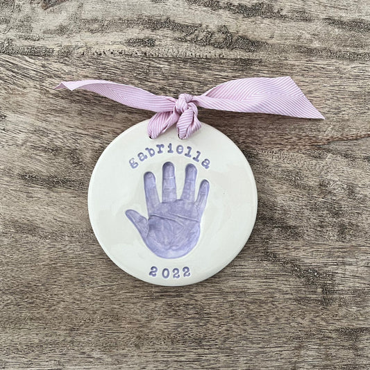 Round ceramic ornament with a lavender baby handprint, personalized with the name Gabriella and year 2022, finished with a pink ribbon.
