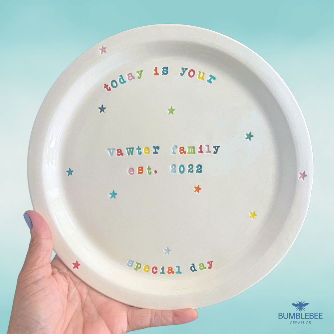 White ceramic plate with colorful text reading “today is your special day,” featuring a family name and “est. 2022” in the center, surrounded by small multicolored star accents, held against a soft blue background.
