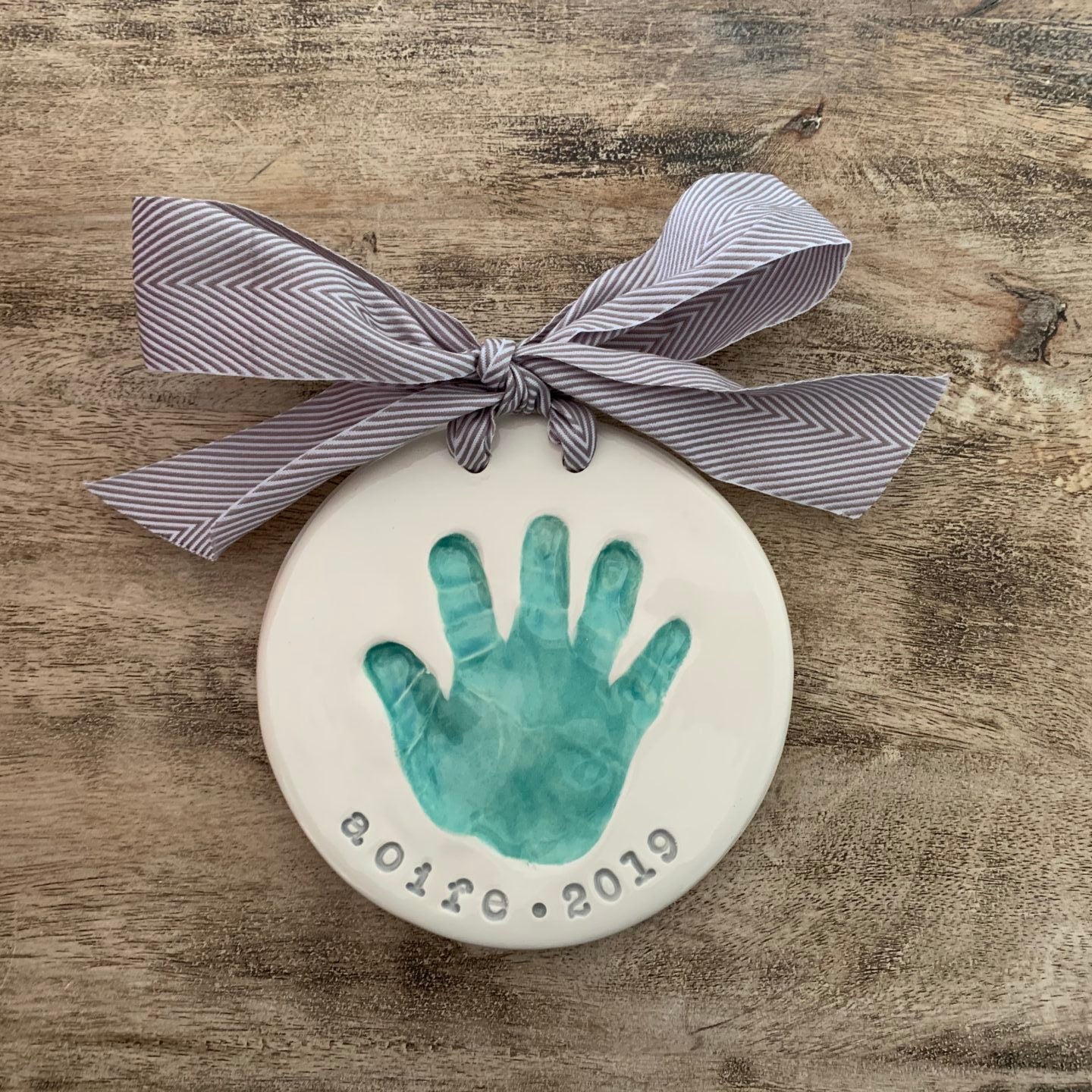 Round ceramic keepsake with a teal baby handprint and the name Aoife with year 2019 stamped below, finished with a gray herringbone ribbon.